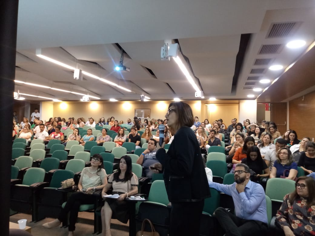 Mulher fala ao microfone para pessoas sentadas em auditório