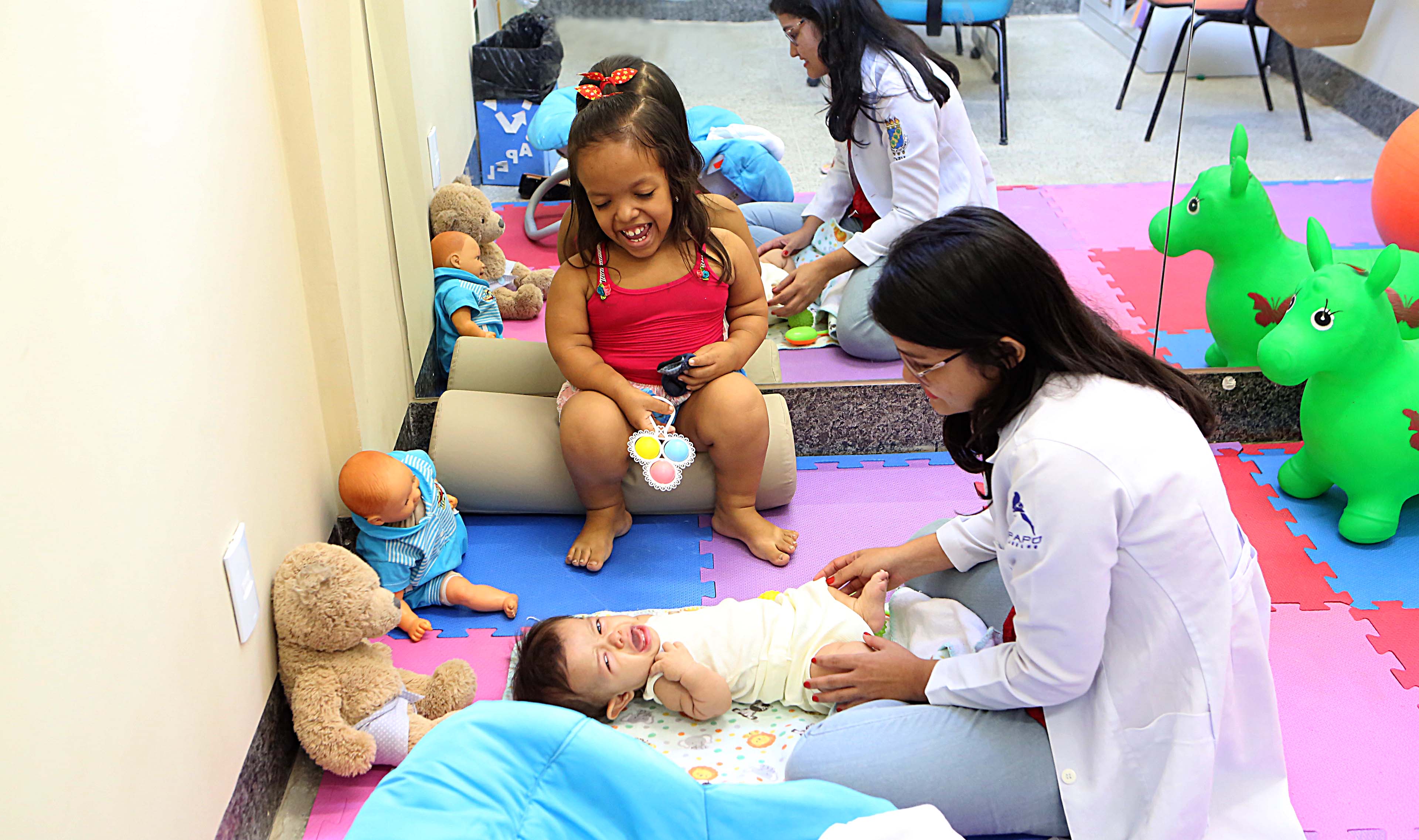 Fisioterapeuta atendendo bebê em tapete no chão com menina assistindo