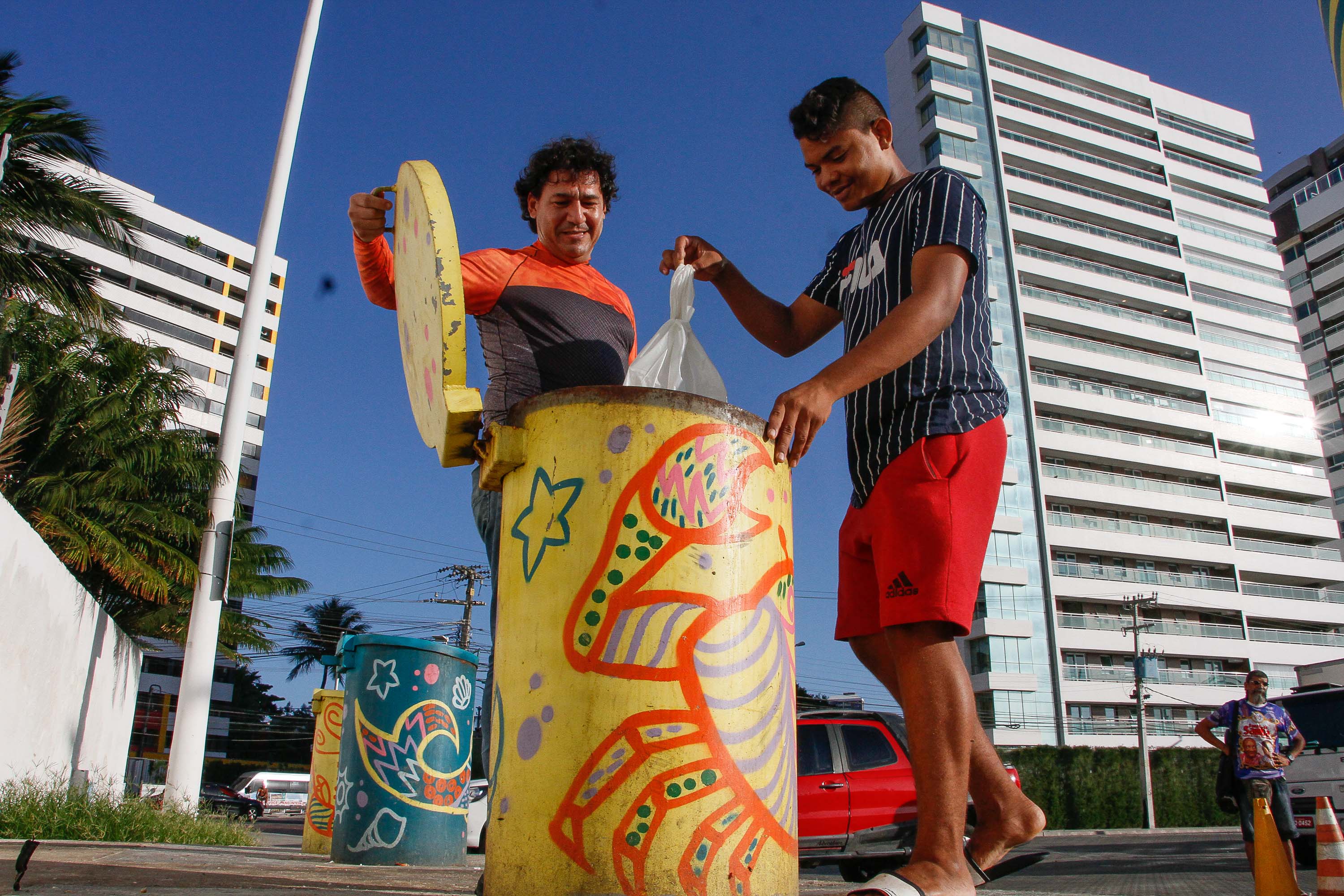 Dois homens colocando lixo em lixeira subterrânea com prédios ao fundo