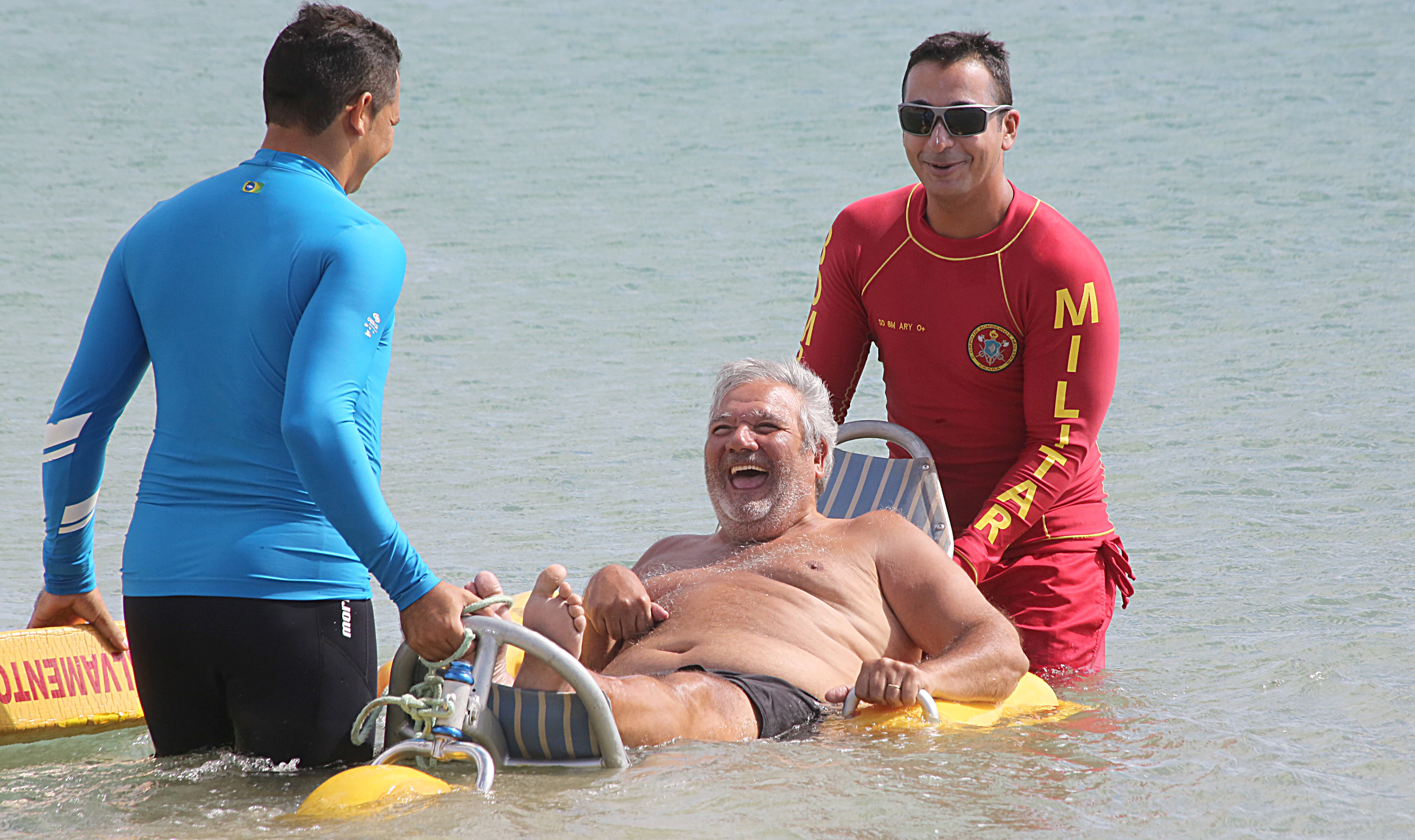 Senhor tomando banho de mar em cadeira acessível ao lado de ajudantes