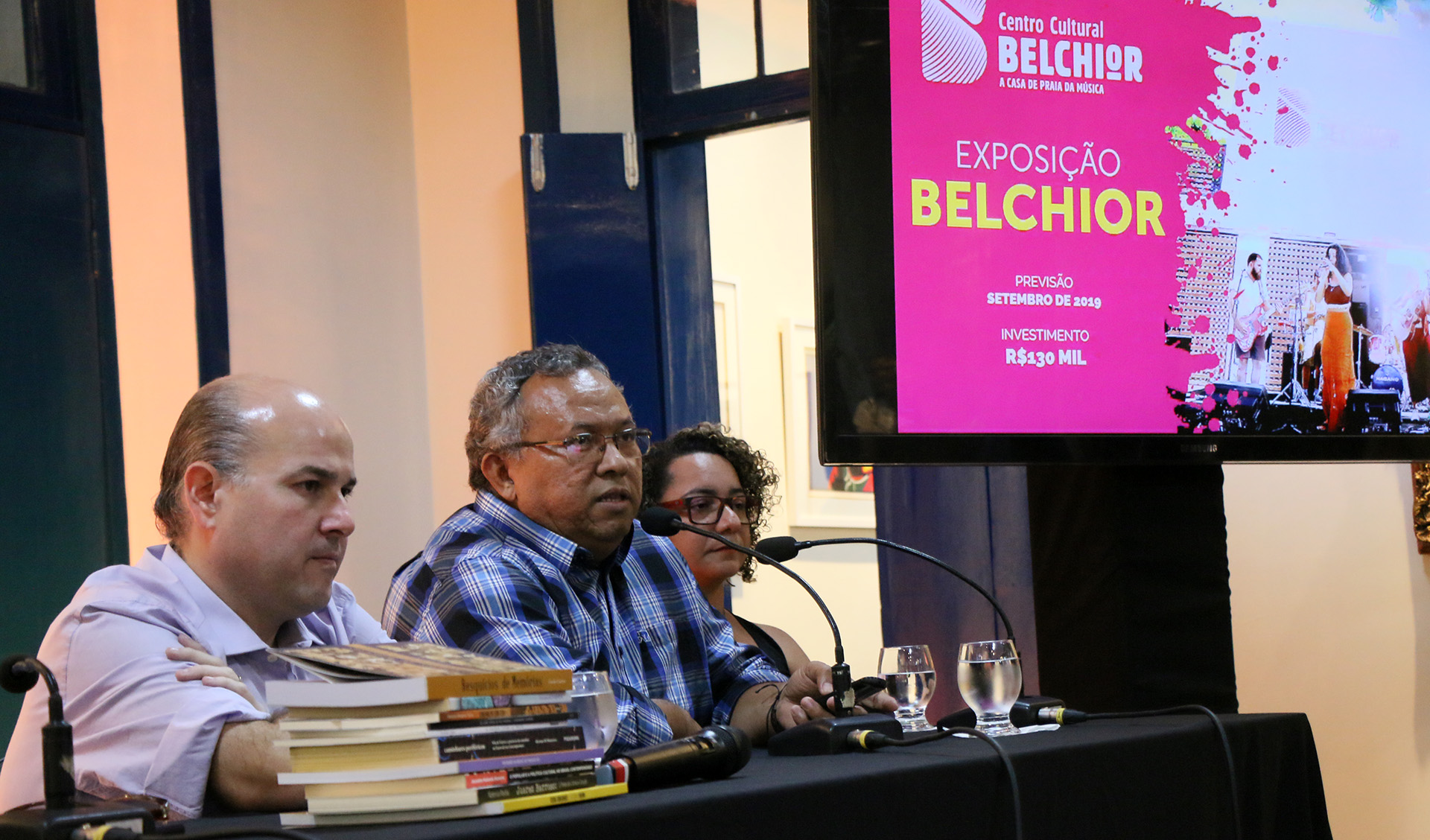 Prefeito, homem e mulher sentados de perfil com tela ao fundo exibindo apresentação sobre a Exposição Belchior