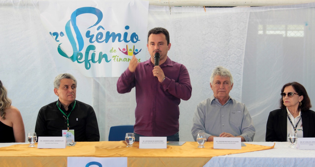 Secretario adjunto da Educação em pé falando ao microfone e pessoas ao lado sentadas em mesa