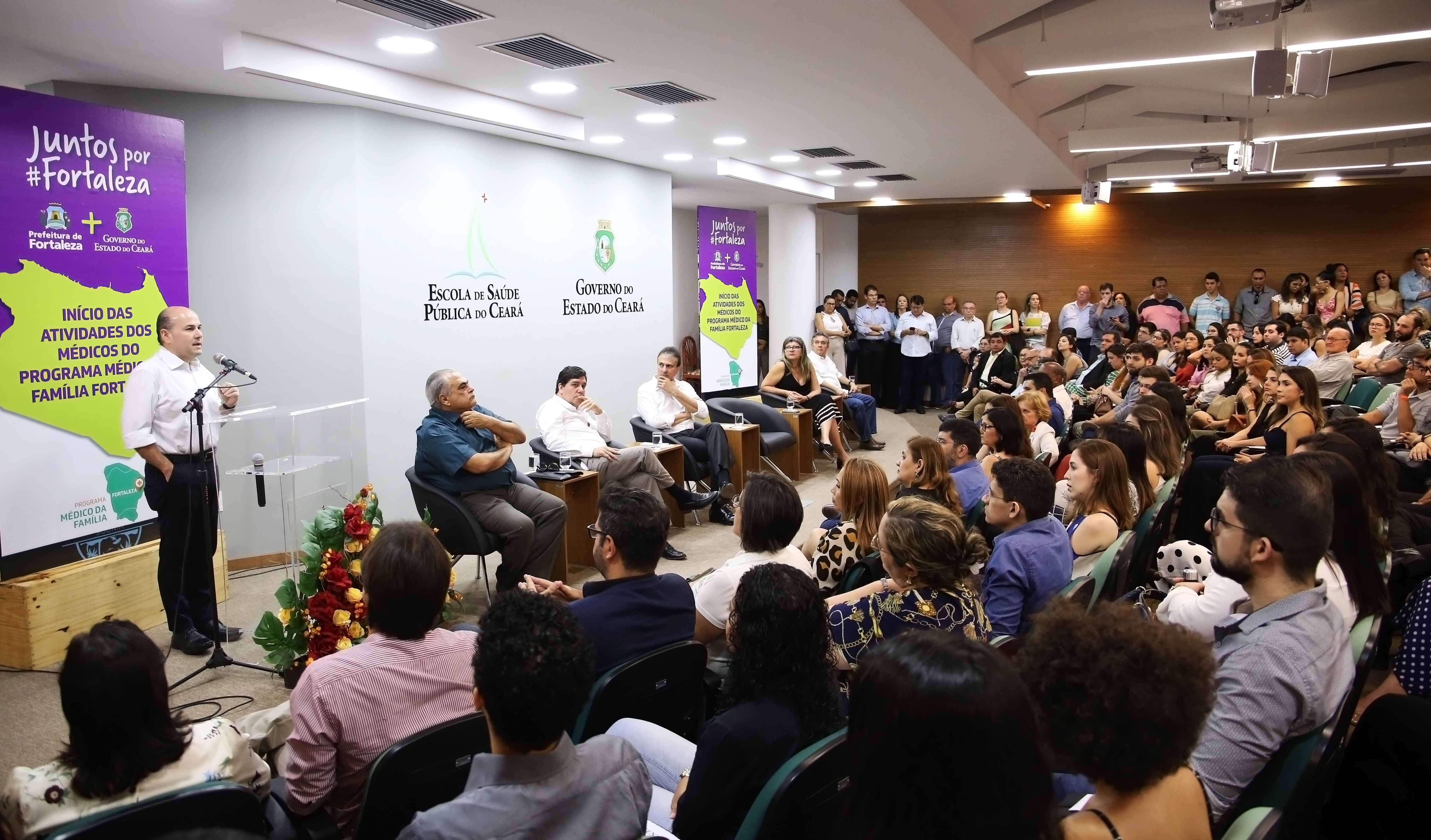 Prefeito falando em púlpito em cima de palco com pessoas sentadas ao fundo e público assistindo