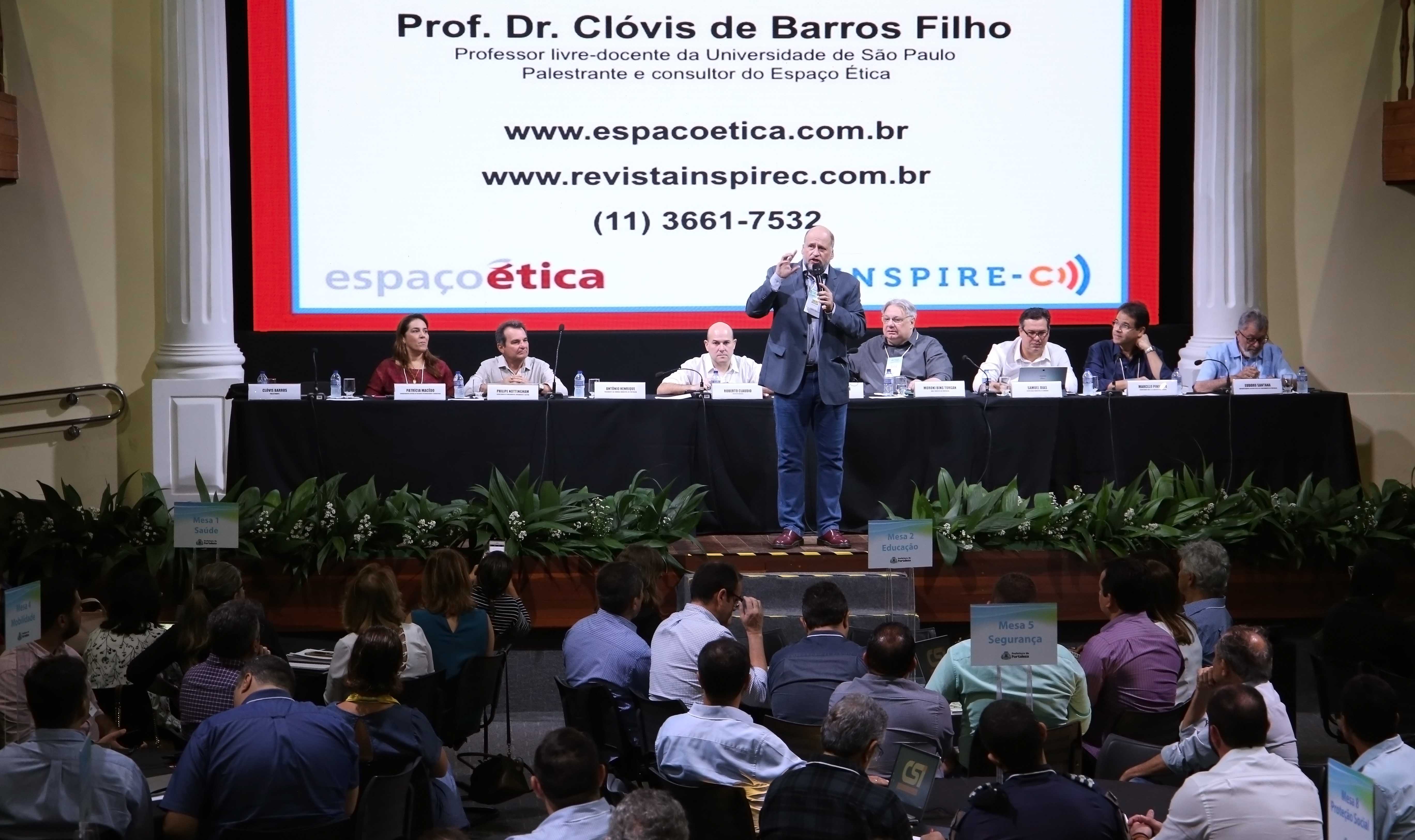 Homem falando ao microfone em cima do palco com pessoas em mesa atrás e outras embaixo assistindo