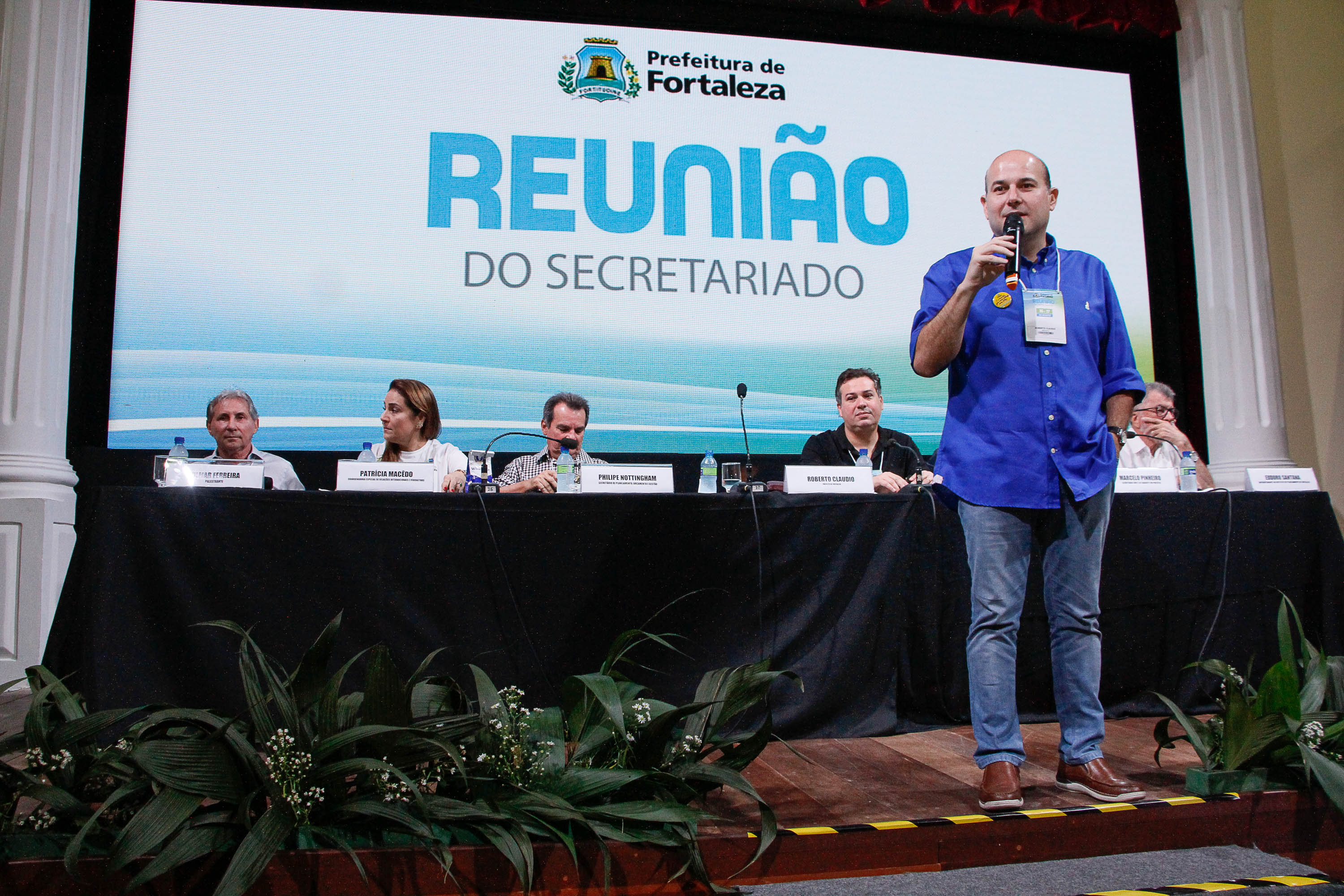 prefeito em pé falando ao microfone com telão e mesa com secretários ao fundo