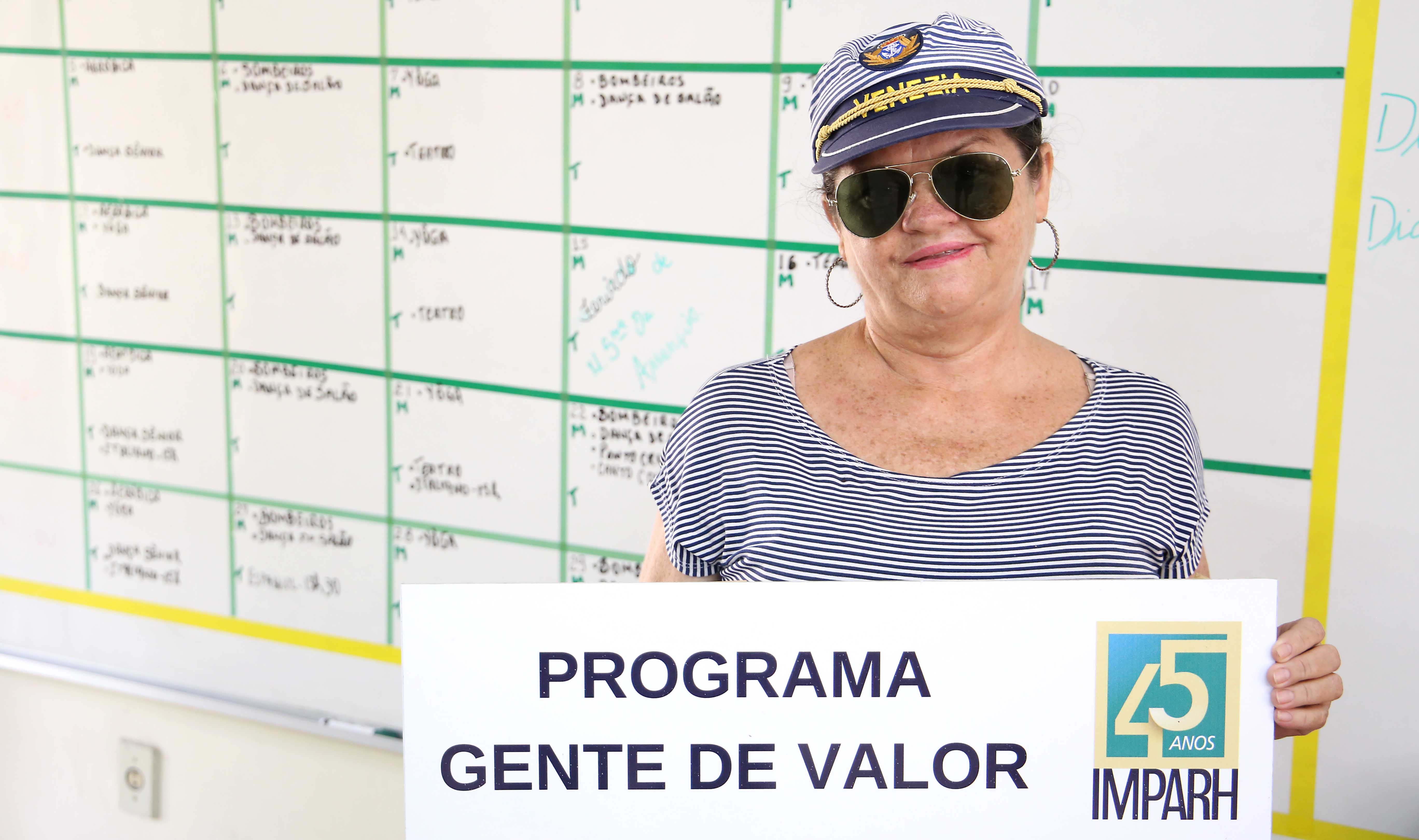 Senhora de óculos escuros e chapeu de capitão posando pra foto e segurando placa escrito programa gente de valor