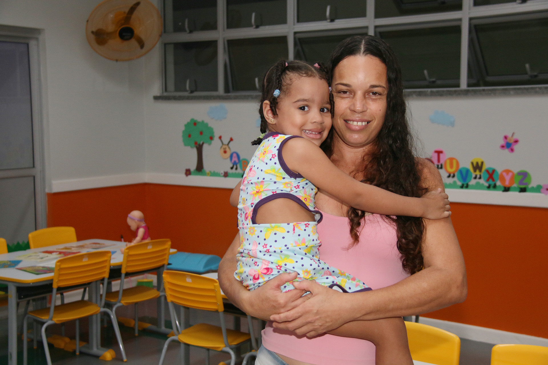 mãe segura filha no colo, as duas sorriem para câmera