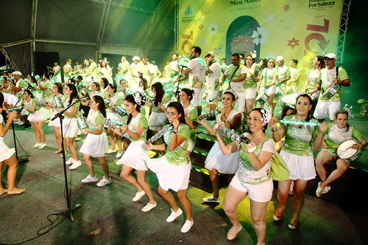 Homens e mulheres membros do Bloco Camaleões do Vila utilizam instrumentos de percussão em cima de um palco na Praia de Iracema