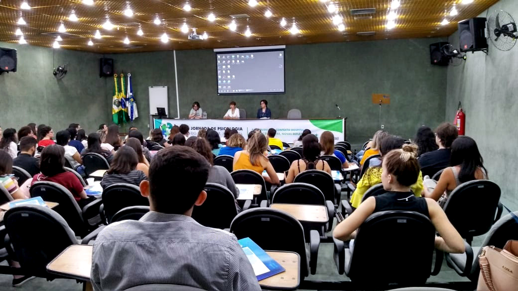 Pessoas sentadas à mesa em cima de palco com plateia assistindo