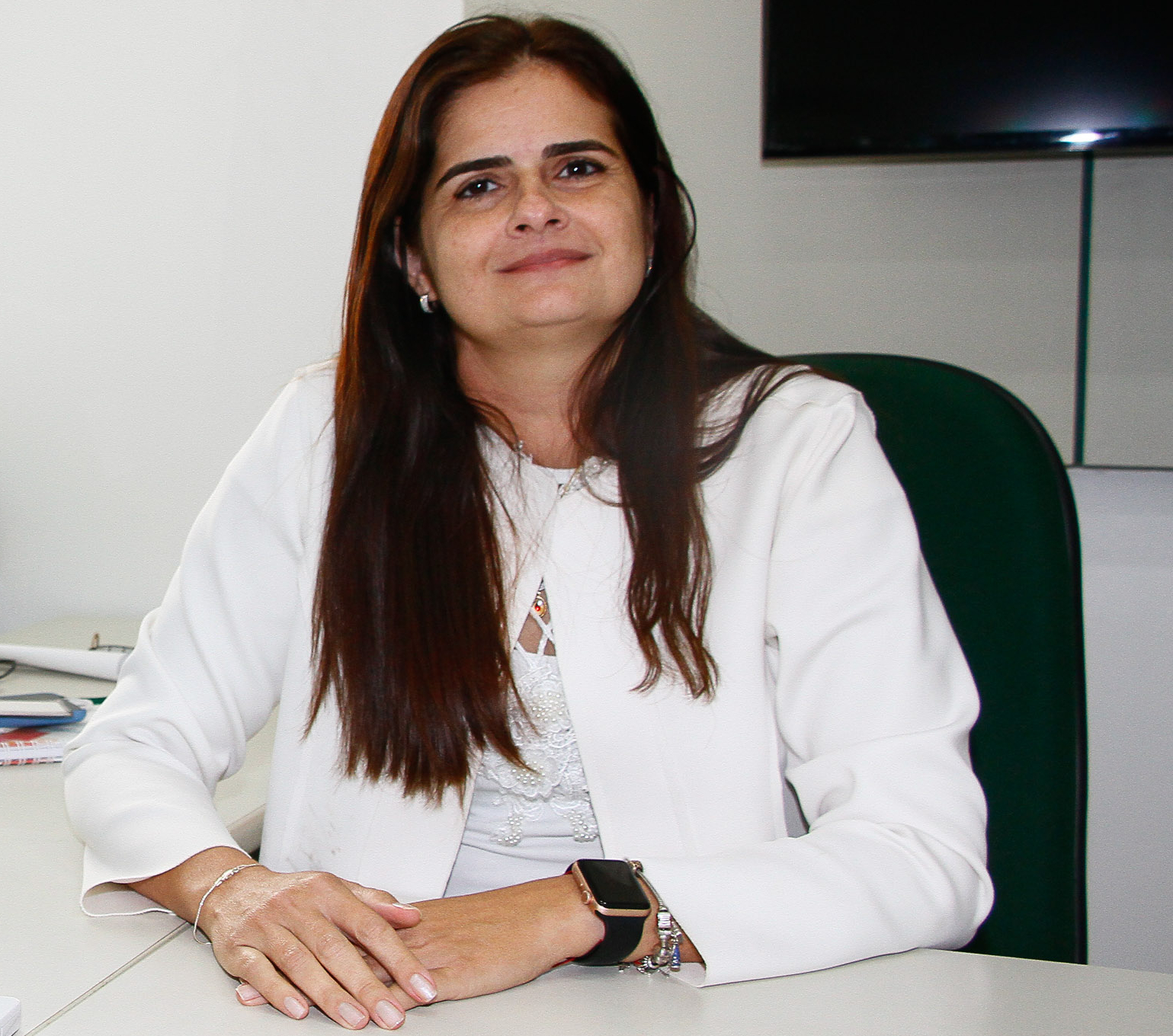 Flávia Teixeira sentada com braços cruzados apoiados em mesa e sorrindo para a foto