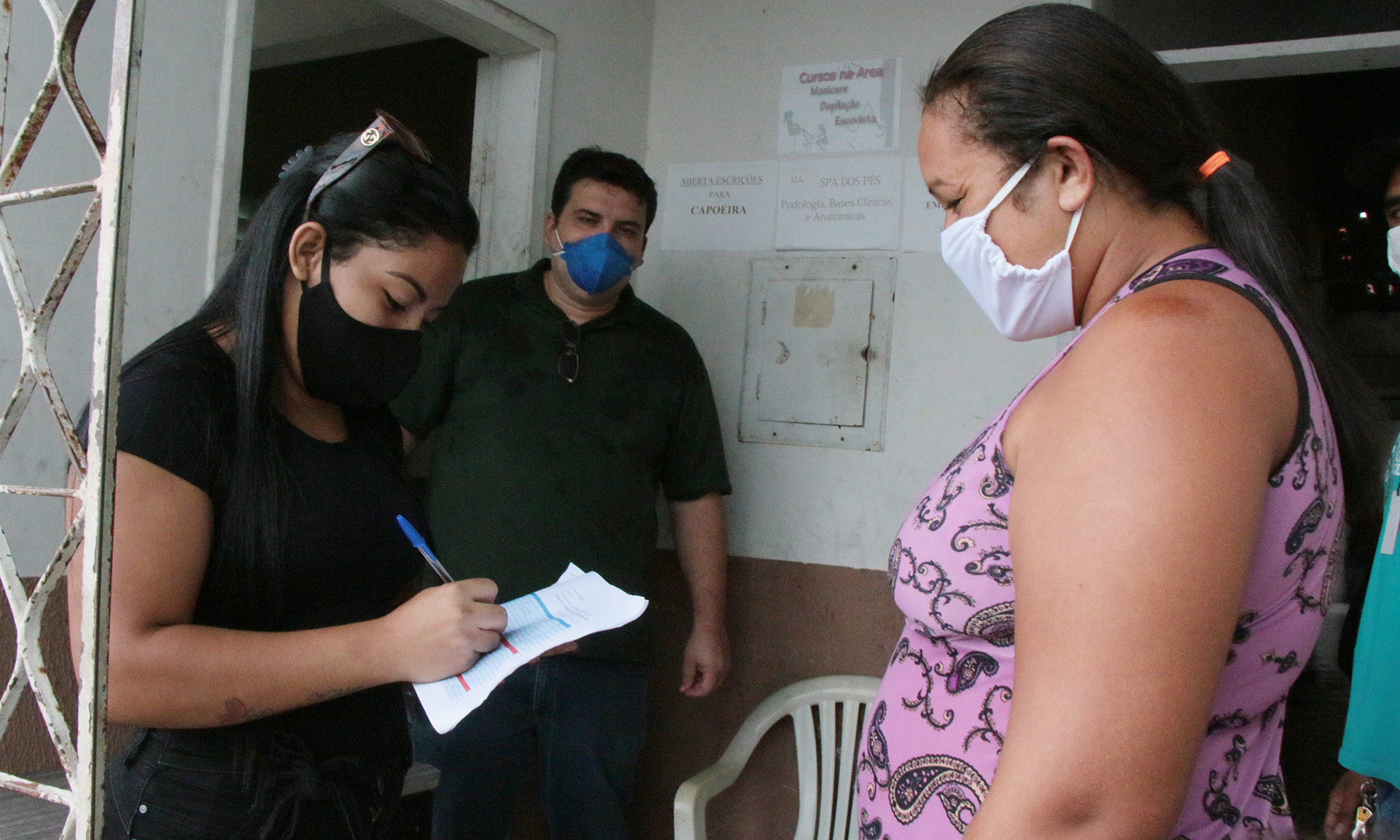 mulher de máscara escrevendo em papel, em frente a outra mulher de máscara olhando pra ela e homem de máscara ao fundo