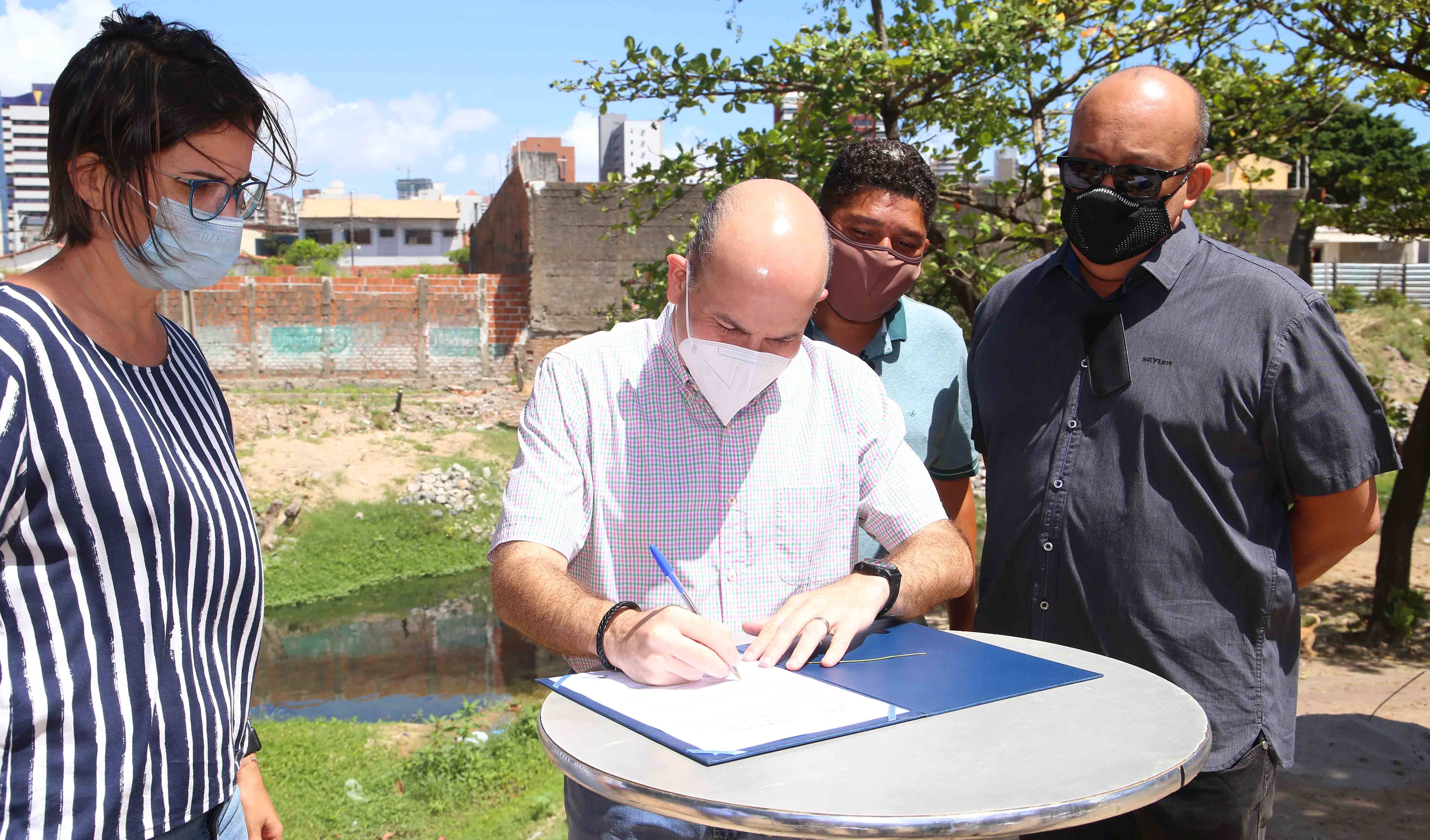 prefeito assinando documento em cima de mesa com riacho maceió ao fundo, secretária Manuela e Paulinho Leme em pé ao lado olhando. todos usam máscara