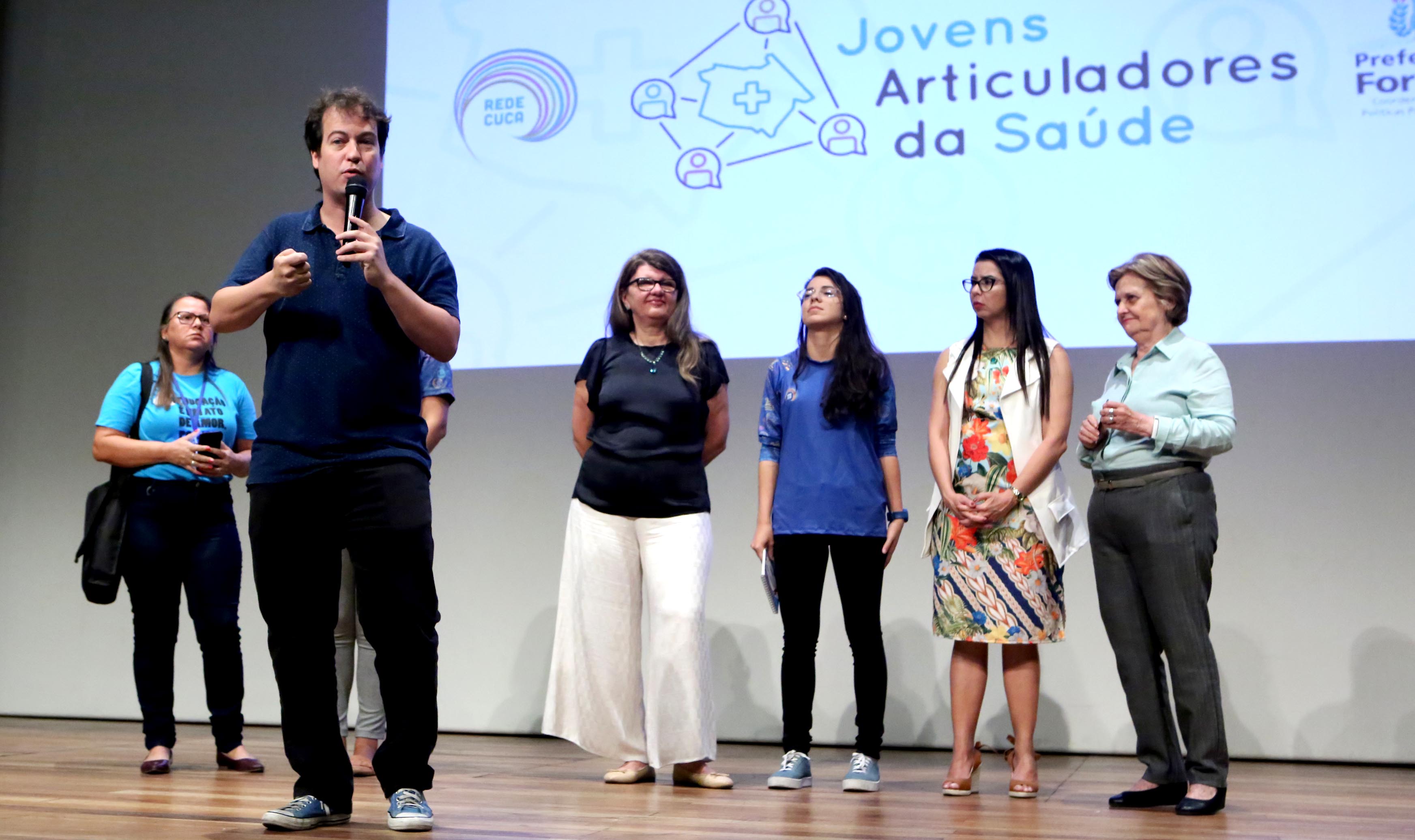 coordenador de juventude em palco falando ao microfone ao lado de mulheres