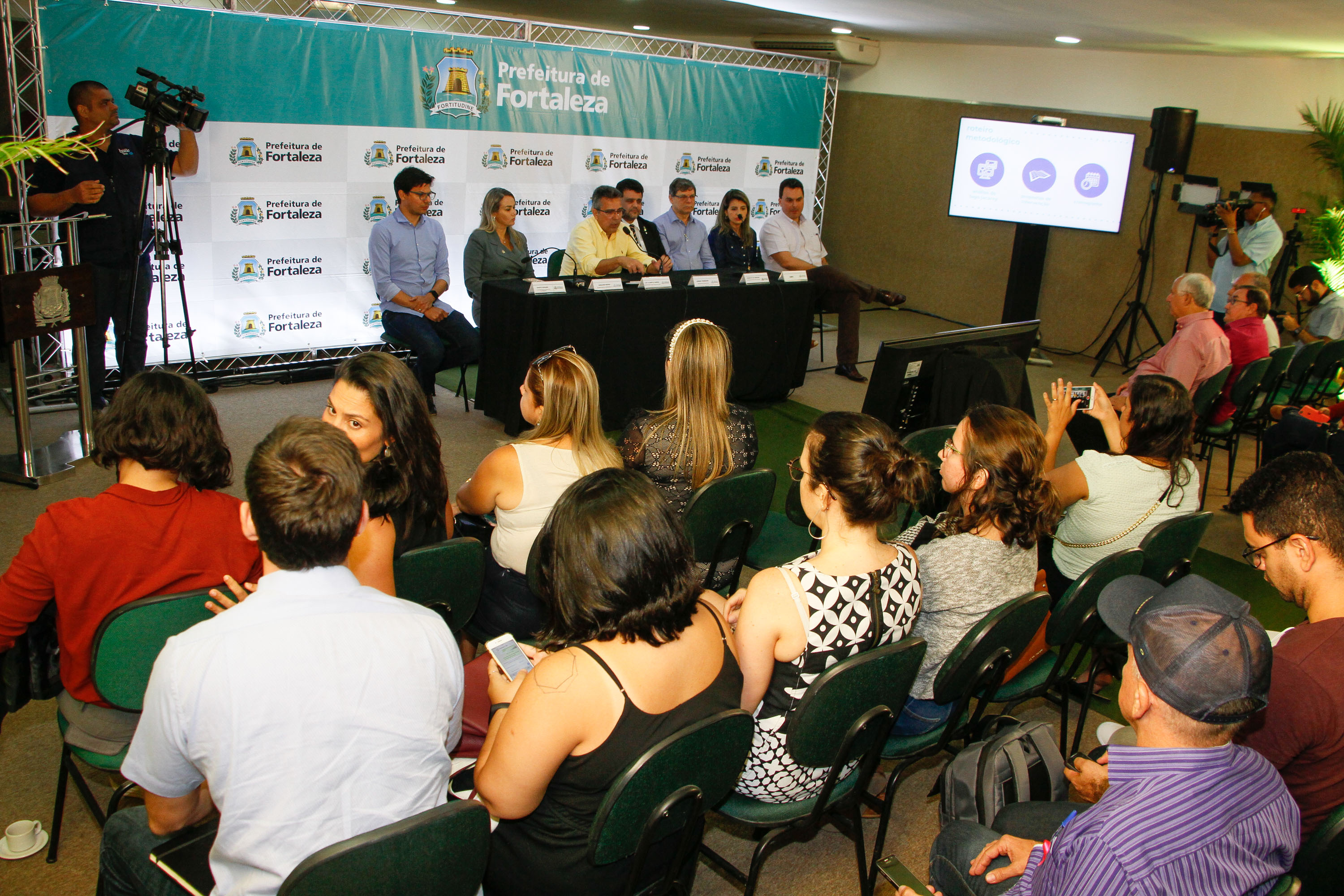 pessoas sentadas em mesa falando ao microfone para plateia sentada à frente