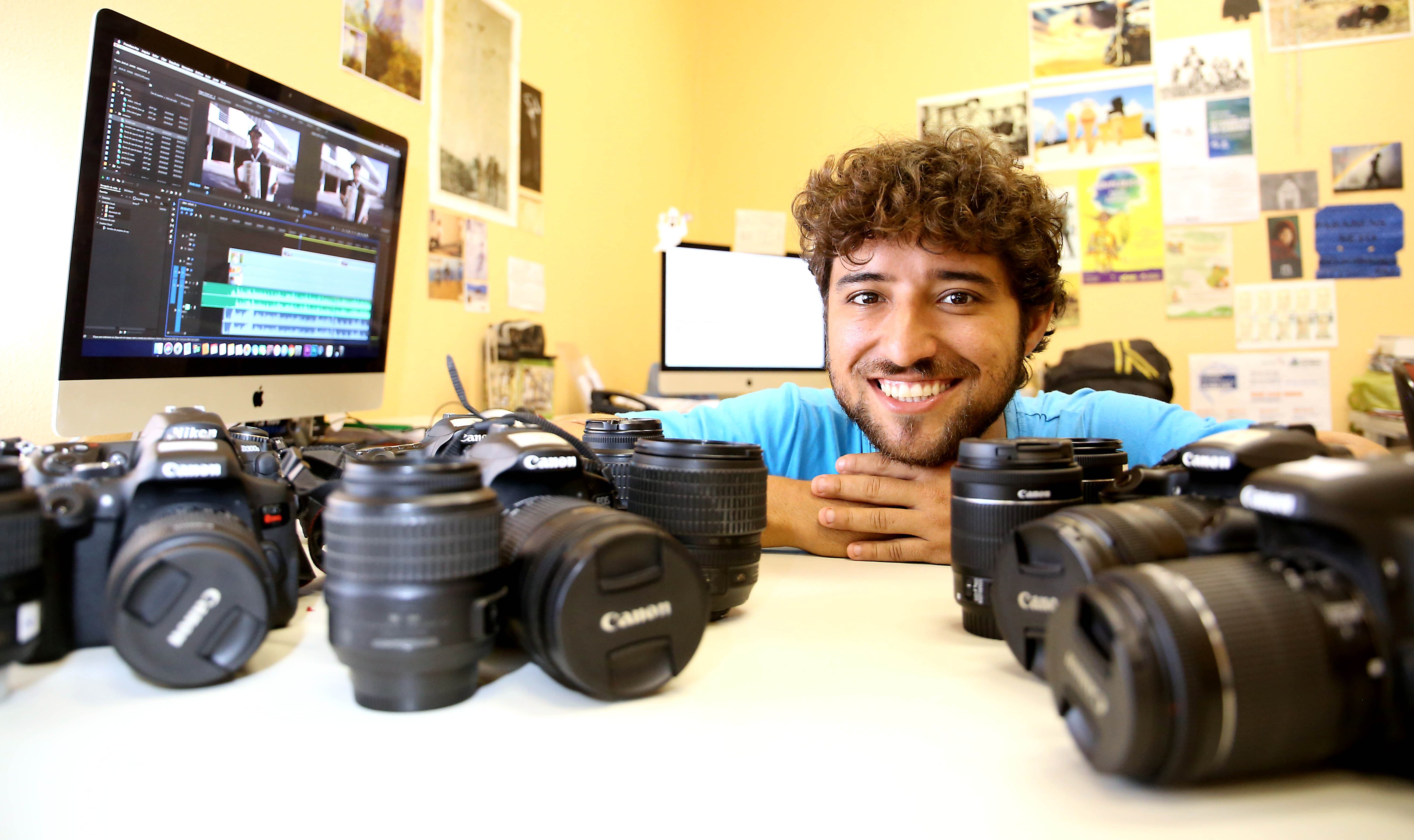 rapaz sorrindo sentado à mesa com várias lentes fotográficas em cima