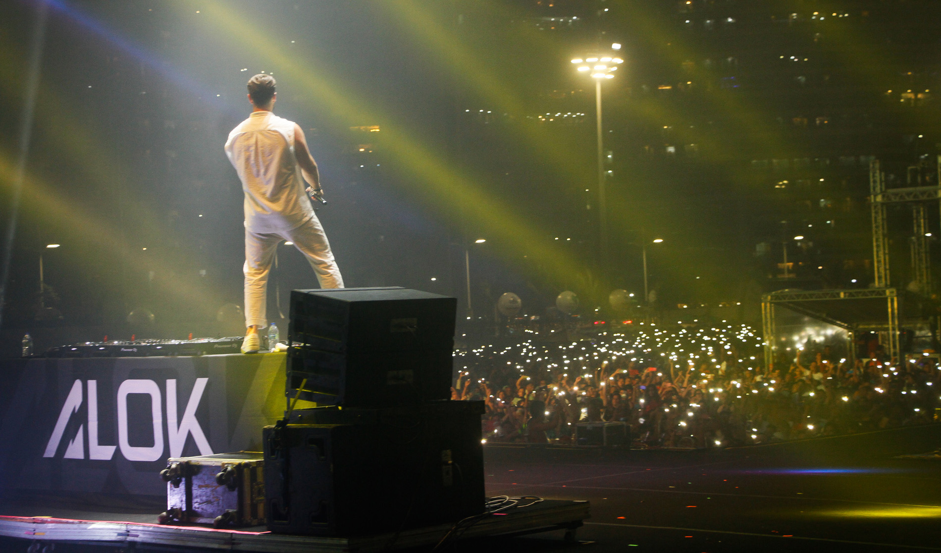 dj em cima de palco com braços pra cima e público em frente assistindo