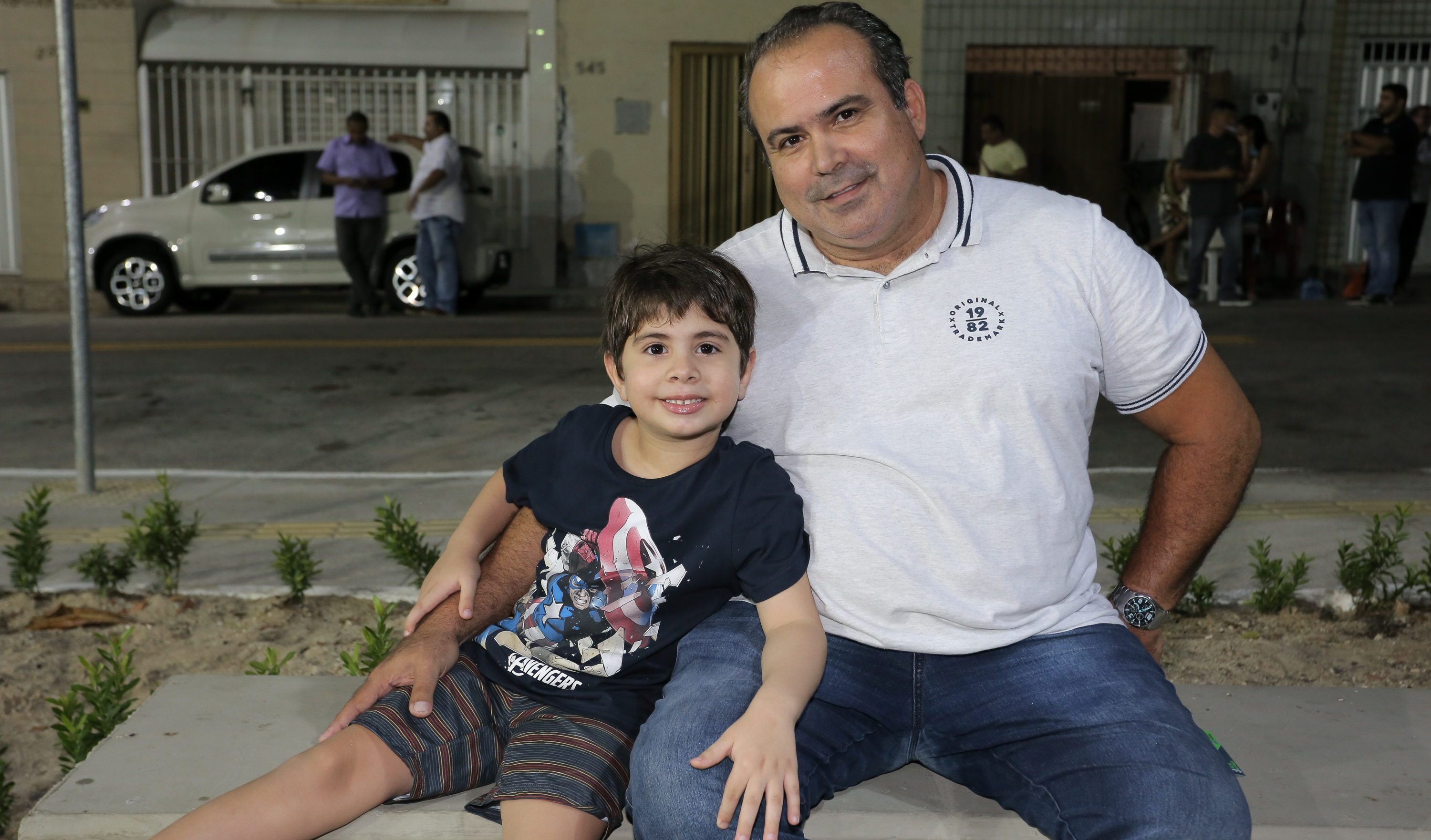 homem e menino sentados lado a lado em banco de praça sorrindo para a foto