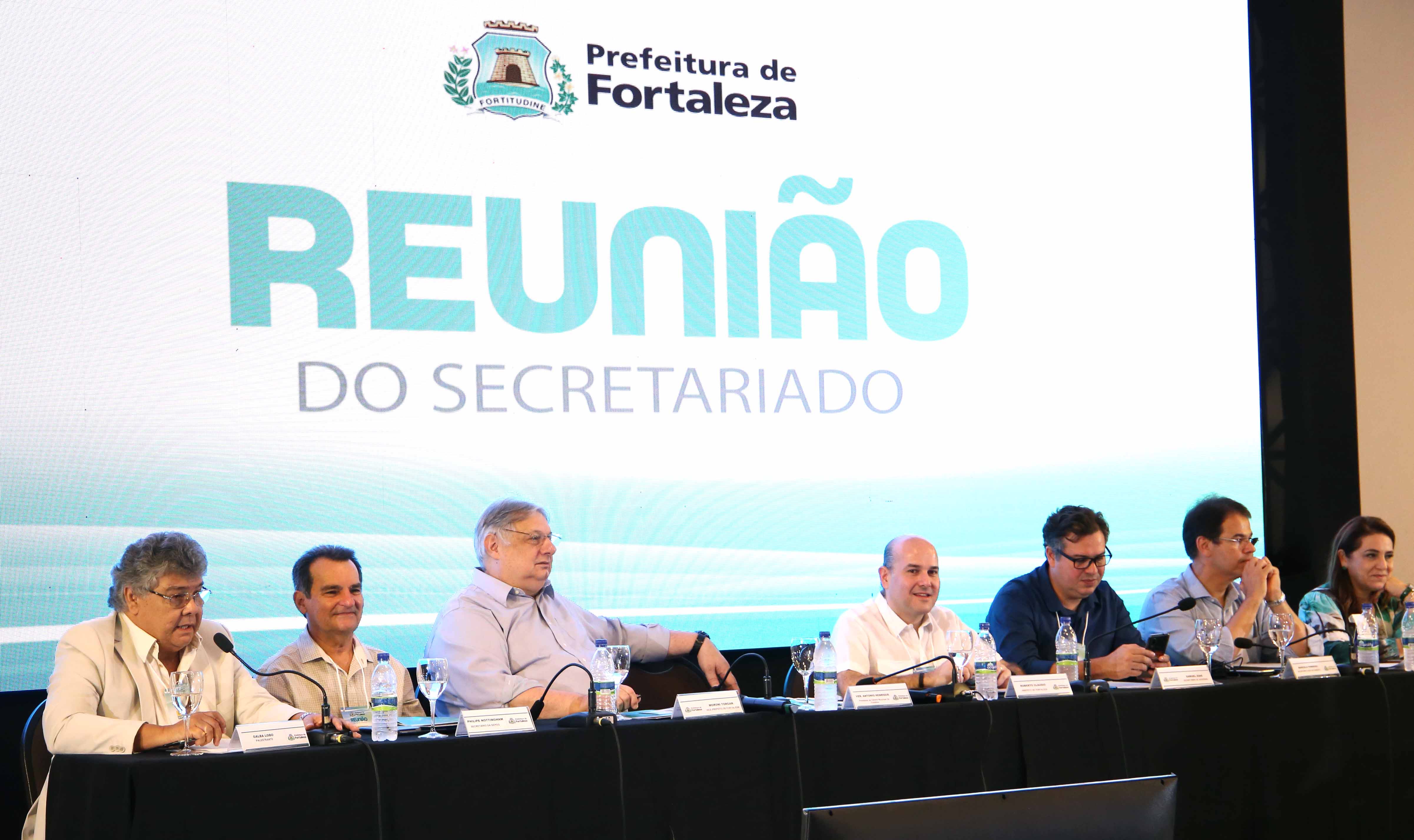 prefeito sentado à mesa em cima de palco ao lado de secretários