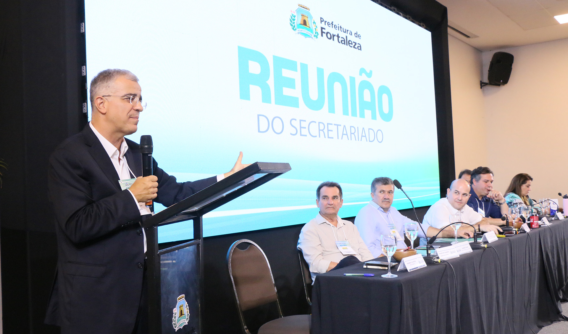 senhor falando ao microfone ao lado de mesa com prefeito e secretários sentados assistindo