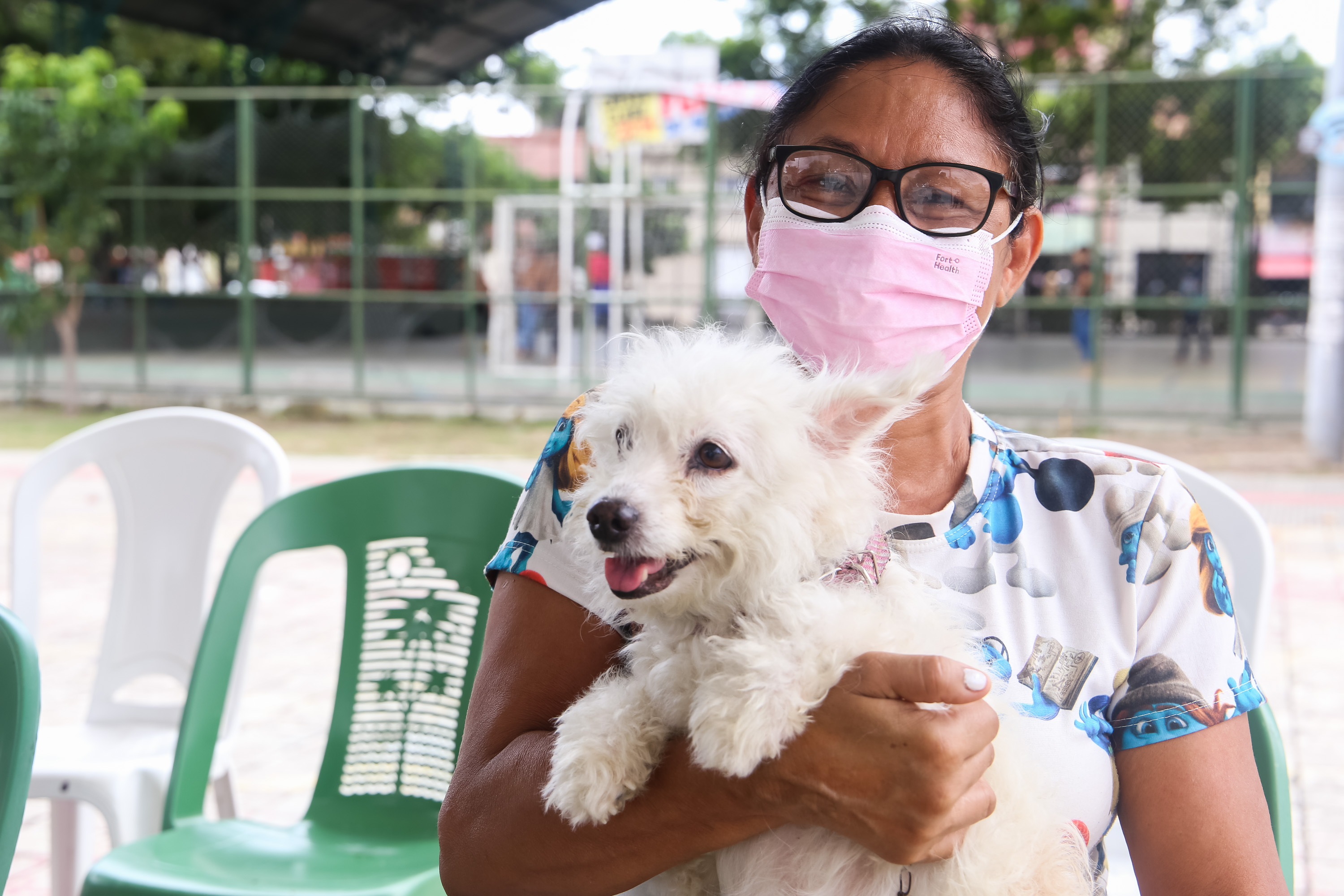 Mulher de máscara e com blusa cor de rosa segura cachorro nos braços. 