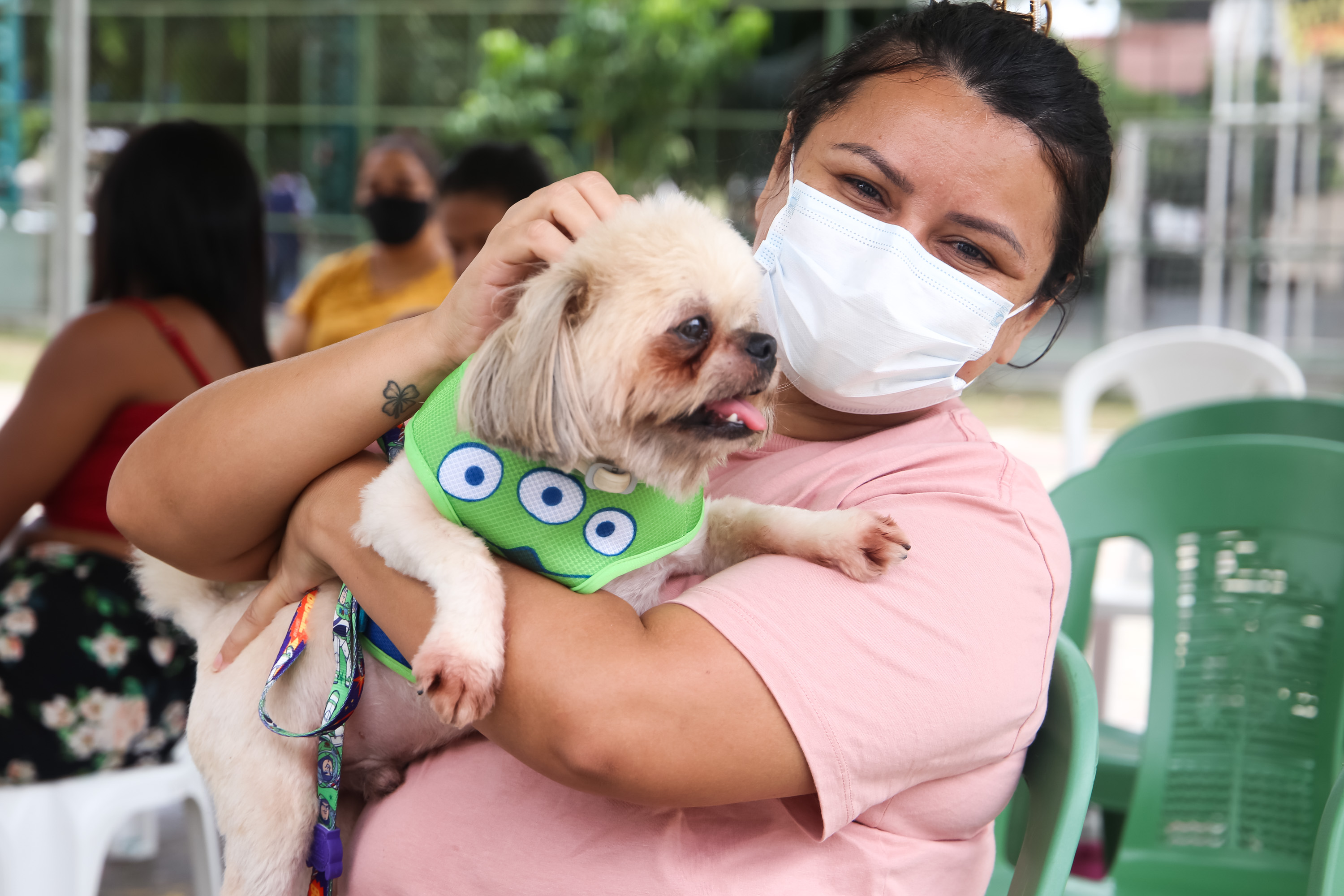 Mulher de máscara e com blusa cor de rosa segura cachorro nos braços. 