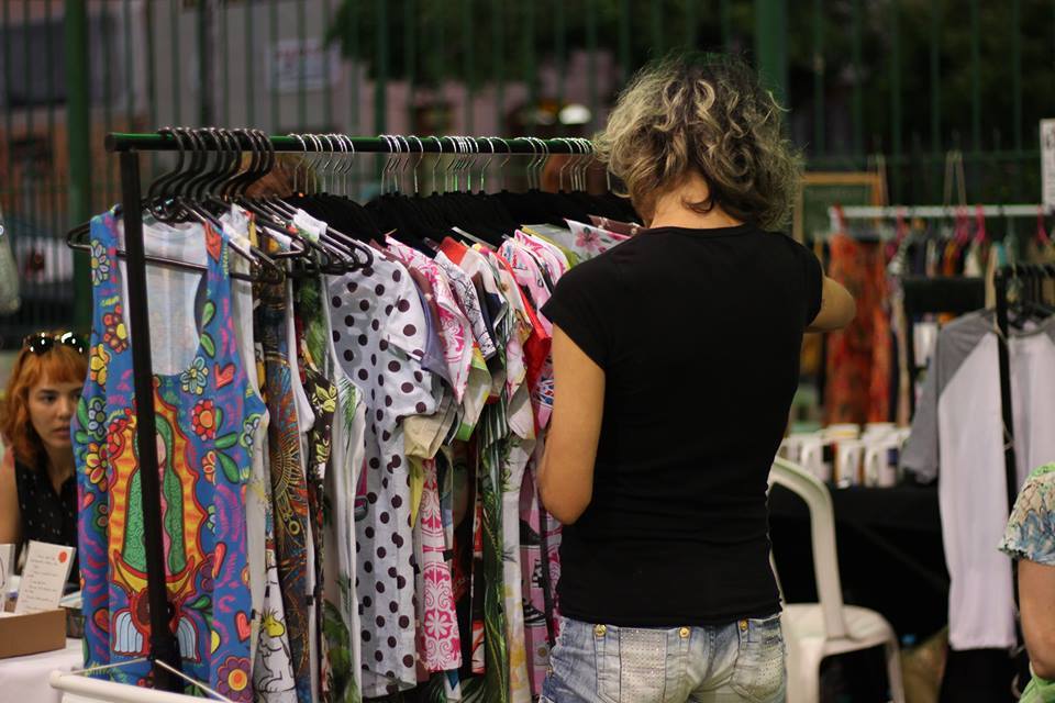 Mulher loira, de costas, com camisa preta olhando camisas penduras em arara