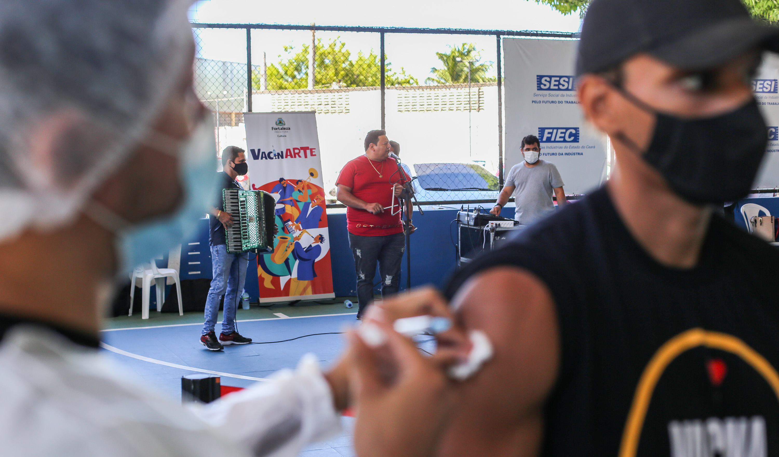 Homem recebendo vacina. Ao fundo, banda tocando.