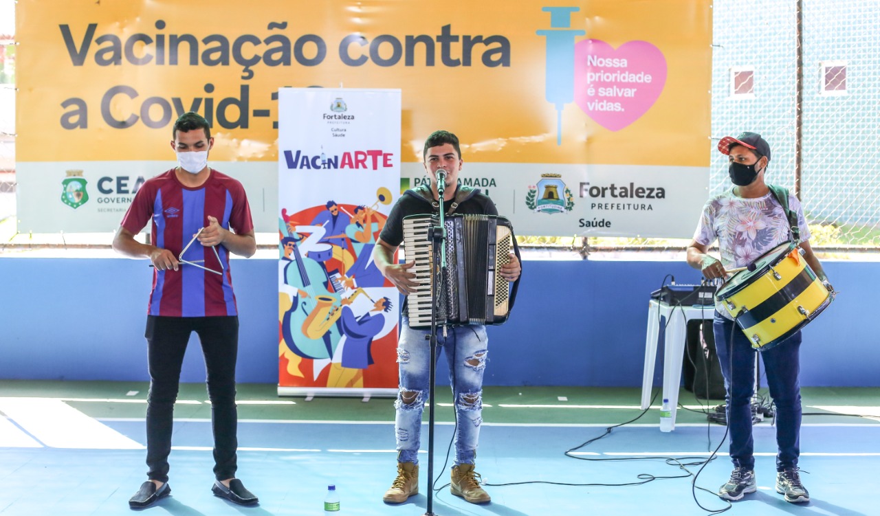 Trio de forró tocando os instrumentos Triângulo, Bumbo e Sanfona, em ginásio, durante vacinação. Ao fundo, a peça gráfica do Vacinarte