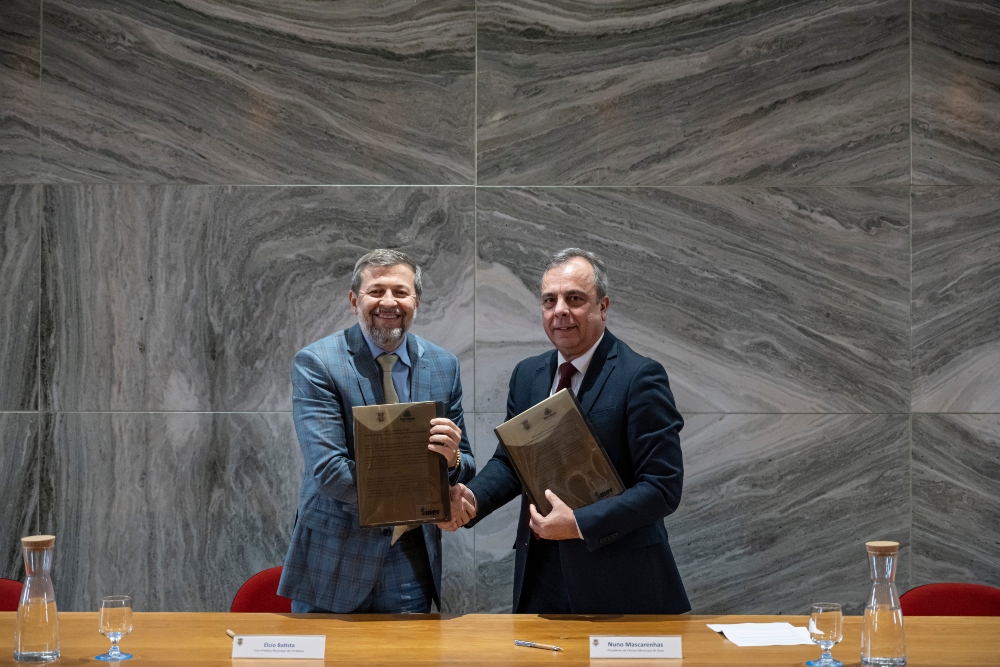 Registro fotográfico mostra, à esquerda, o vice-prefeito de Fortaleza, Élcio Batista; e, à direita, o presidente da Câmara Municipal de Sines, Nunu Mascarenhas. Ambos estão em pé, atrás de uma bancada de madeira, e seguram os documentos que simbolizam o acordo firmado. Élcio usa um paletó de cor azul-claro, quadriculado, e Nuno usa um terno de cor azul-escuro. Atrás dos dois, há uma parede de mármore, cinza. Em cima da bancada, há garrafas e copos com água.