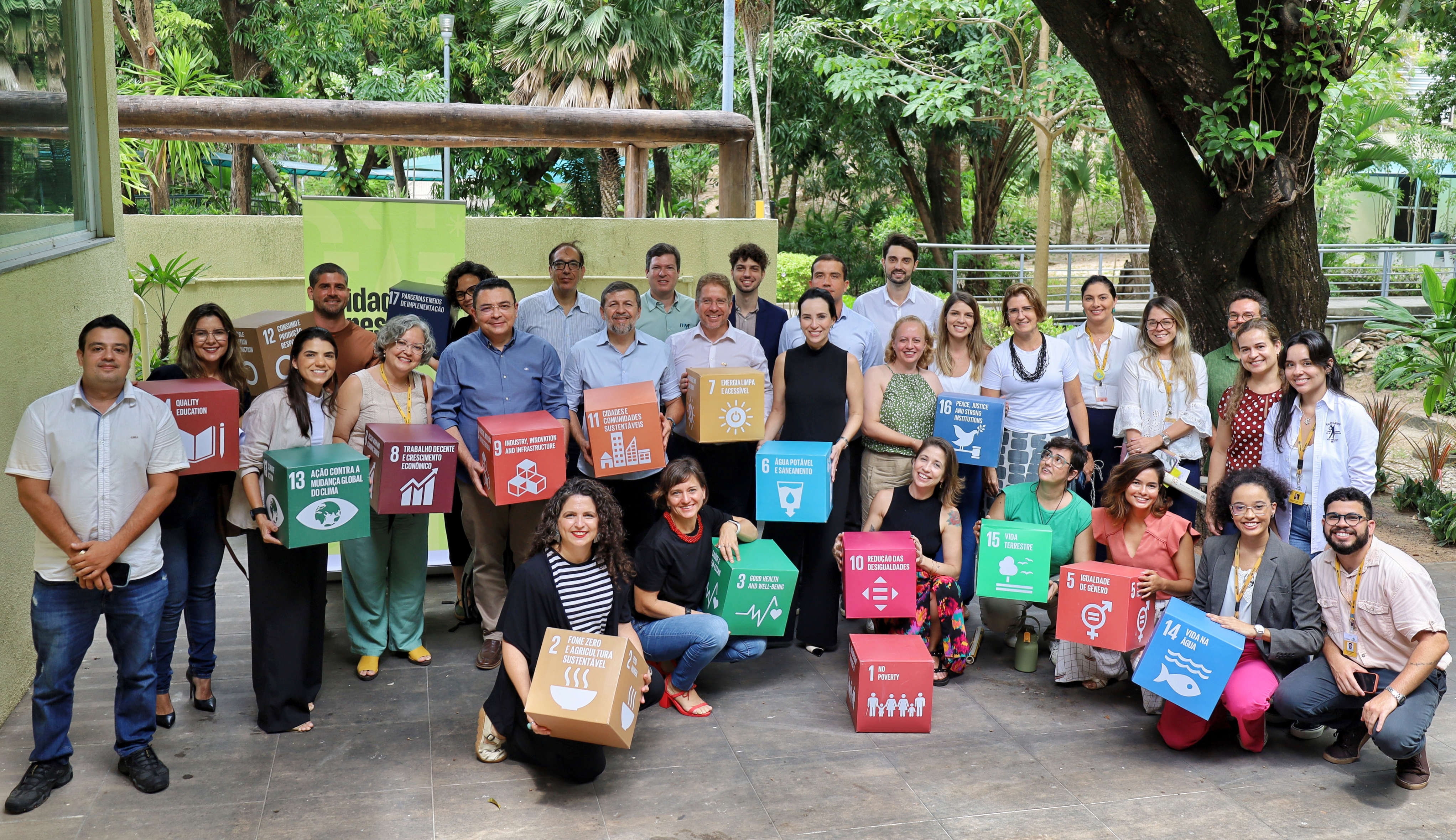 A fotografia mostra representantes de diversas instituições posando para a imagem. Alguns estão em pé, outros estão de cócoras. Alguns seguram dados coloridos que representam os Objetivos de Desenvolvimento Sustentável da ONU. Ao fundo, está o bosque do Paço Municipal de Fortaleza.  